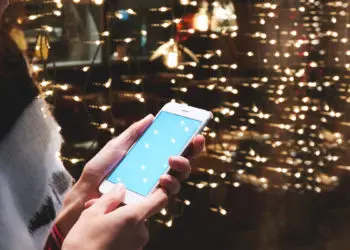 Closely of woman is reading text message in social network via smart phone, while is standing outdoors against garlands lights. Hipster girl is reading information on web page via her cell telephone