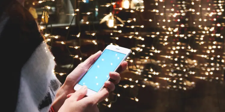 Closely of woman is reading text message in social network via smart phone, while is standing outdoors against garlands lights. Hipster girl is reading information on web page via her cell telephone